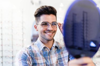 Yakışıklı genç adam gözlük çerçevesini optik mağazada seçiyor..