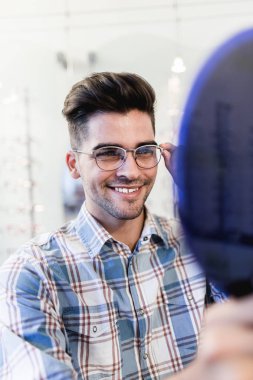 Yakışıklı genç adam gözlük çerçevesini optik mağazada seçiyor..
