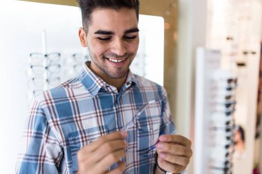 Yakışıklı genç adam gözlük çerçevesini optik mağazada seçiyor..