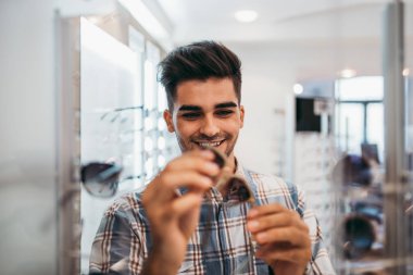 Yakışıklı genç adam gözlük çerçevesini optik mağazada seçiyor..
