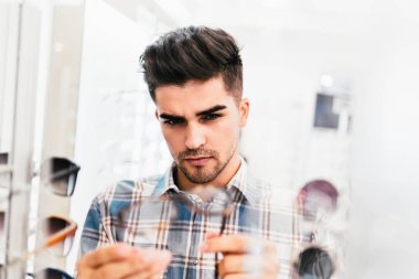 Yakışıklı genç adam gözlük çerçevesini optik mağazada seçiyor..