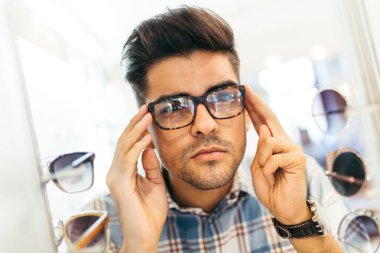 Yakışıklı genç adam gözlük çerçevesini optik mağazada seçiyor..