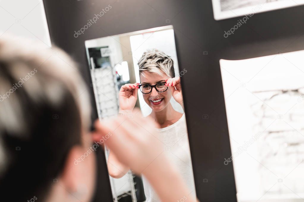 Beautiful middle age woman choosing a glasses in optician store.