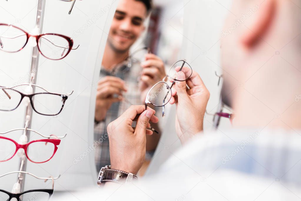 Handsome young man choosing eyeglasses frame in optical store.