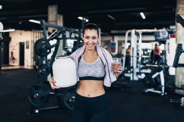 Genç çekici kadın başarılı egzersiz poz ve modern fitness spor salonunda gülümseyen sonra büyük beyaz protein kavanoz ve diğer taraftan içme saman ile sallamak cam tutarken.