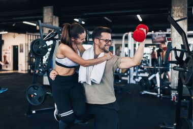 Genç çekici çift gülümseyen ve modern fitness spor salonunda eğlenceli çalışma. Adam bir el ve kırmızı dumbbell ile başka bir kadın tutan.
