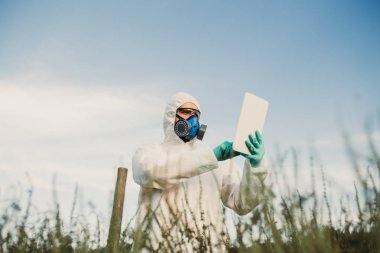 Ot kontrolü. Endüstriyel Tarım araştırması. Koruyucu süitte dijital tableti olan ve alanda ot örnekleri alan bir maske olan adam. Güneşli gün doğal sert ışık. 