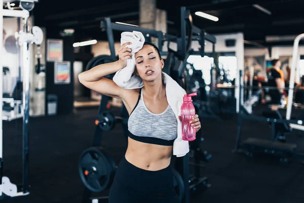 Young exhausted woman in fitness gym after successful workout wipes sweat from the forehead with white towel while holding water bottle in other arm.