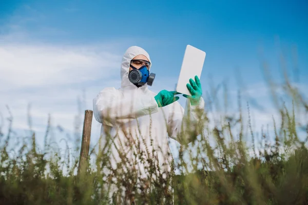 Weed control. Industrial agriculture researching. Man with digital tablet in protective suite and mask taking weed samples in the field. Natural hard light on sunny day.