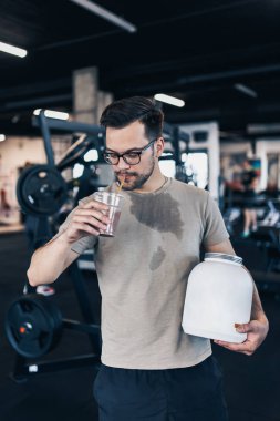 Genç ve çekici bir adam, modern spor salonunda başarılı bir idmandan sonra elinde büyük beyaz protein kavanozu ve diğer elinde pipetle içki içen bir adam..