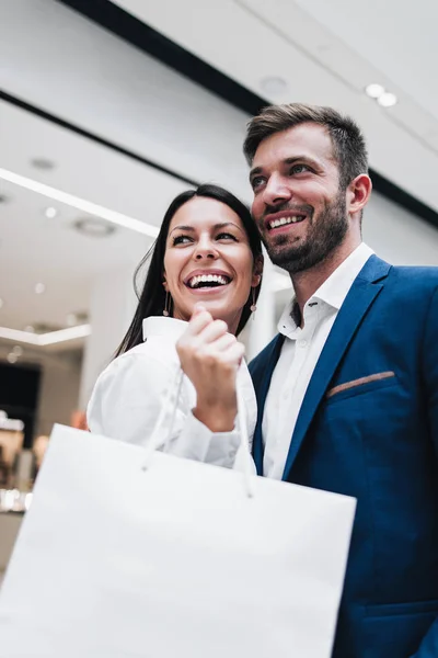 Beautiful couple enjoying in shopping mall or center. They standing in front of huge modern store, smiling and looking at side while holding shopping bags. Shoot from below.
