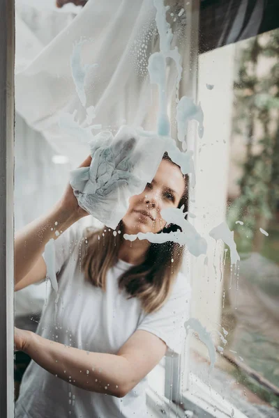 Beautiful and diligent middle age handy woman cleaning windows at her home or apartment and listening to music with headphones. Do it yourself housework concept.