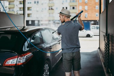 Gündüz vakti araba yıkama istasyonunda yüksek basınçlı su kullanan şapkalı genç bir adam arabasını yıkıyor..