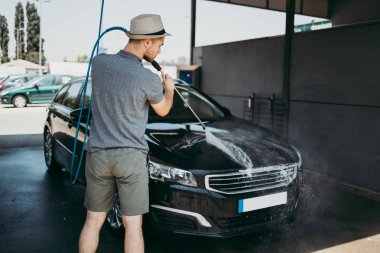 Gündüz vakti araba yıkama istasyonunda yüksek basınçlı su kullanan şapkalı genç bir adam arabasını yıkıyor..