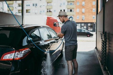 Gündüz vakti araba yıkama istasyonunda yüksek basınçlı su kullanan şapkalı genç bir adam arabasını yıkıyor..