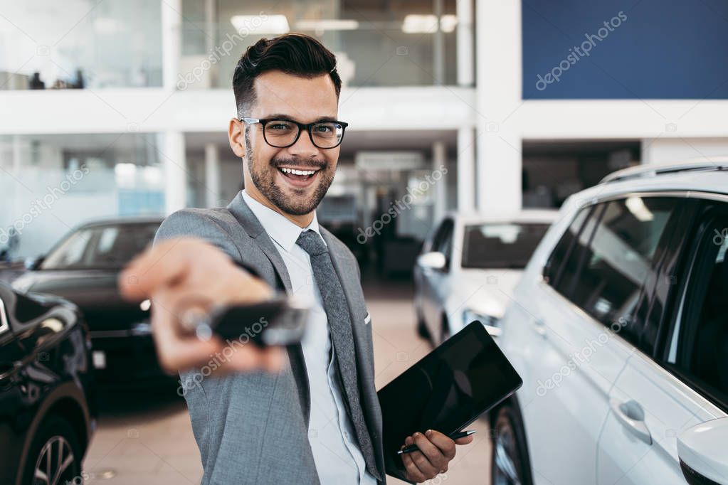 Good looking, smiling salesman poses in a car salon or showroom. He stands with outstretched hand and holds the keys.