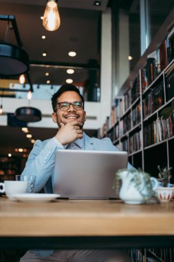 Genç çekici bir adam modern kitapçıda oturuyor, dizüstü bilgisayarını okuyor ve kullanıyor.