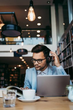 Genç çekici bir adam modern kitapçıda oturmuş, kulaklıkla müzik dinlerken dizüstü bilgisayarını ya da bilgisayarını okuyor ve kullanıyor.