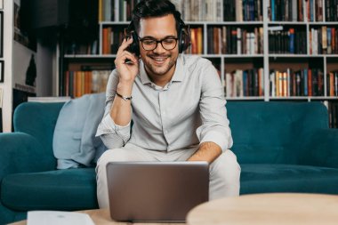 Genç, hoş görünümlü sanatçı stüdyo veya oturma odasında oturuyor, müzik dinliyor, dizüstü bilgisayarında çalışıyor, gülümsüyor ve bir sonraki başarılı projesini düşünüyor. Arkasında kitap ya da kütüphane olan duvar. Aşağıdan ateş et.