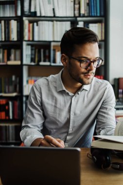 Genç, yakışıklı bir öğrenci ya da yazar pencerenin yanındaki üniversite kütüphanesinde oturuyor ve dizüstü bilgisayarı üzerinde çalışıyor. O ciddi ve yorgun. Güçlü ve derin gölgeleri olan doğal ışık. Arka planda kitap olan duvar.