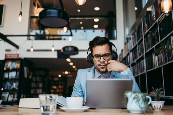 Genç çekici bir adam modern kitapçıda oturmuş, kulaklıkla müzik dinlerken dizüstü bilgisayarını ya da bilgisayarını okuyor ve kullanıyor.