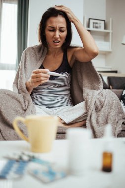 Grip hastası ve yorgun orta yaşlı bir kadın. Evdeki kanepede oturuyor, termometreyi tutuyor ve bej battaniyeye sarılı vücut ısısını ölçüyor..