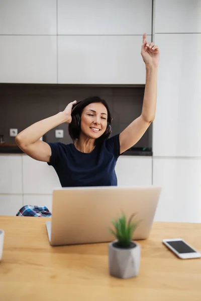 Attractive and happy middle age female freelancer is working at her home. She is using headphones, listening to music and singing. Modern kitchen in background. Freelancing job concept.