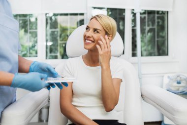 Beautiful and happy blonde woman at beauty medical clinic. She is sitting and talking with female doctor about face esthetics treatment. clipart