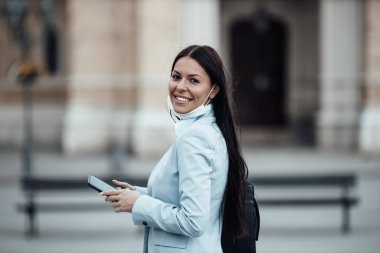 Zarif bir kadın boş bir caddede yürüyor ve kendini tehlikeli grip ya da virüsten korumak için koruyucu maske takıyor. Gülümsüyor ve iyimser. Corona virüsü veya Covid-19 Olumlu kalmaya devam et.