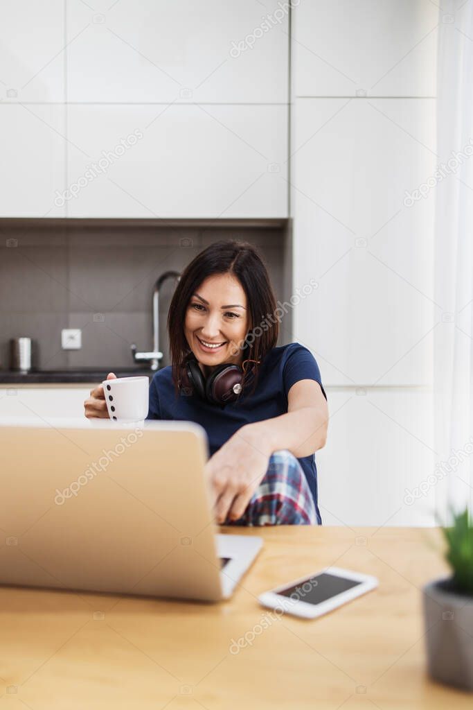 Attractive and happy middle age female freelancer is working and smiling at her home. Modern kitchen in background. Freelancing job concept.