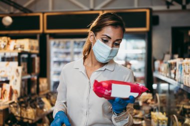 Yüzü koruyucu maskeli ve eldivenli genç ve zarif bir kadın modern bir süpermarkette ya da markette sağlıklı yiyecek ve içecek satın alıyor. Salgın ya da salgın yaşam tarzı ve tüketim kavramı.