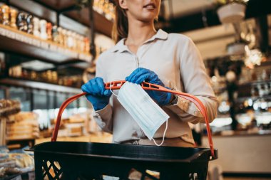 Yüzü koruyucu maskeli ve eldivenli genç ve zarif bir kadın modern bir süpermarkette ya da markette sağlıklı yiyecek ve içecek satın alıyor. Salgın ya da salgın yaşam tarzı ve tüketim konsepti. Maskeye yakın çekim.