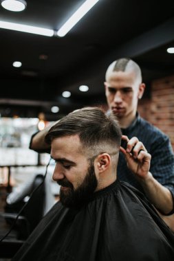 Yakışıklı bir genç adam saç ve sakal stili yaptırıyor ve profesyonel kuaför tarafından giydiriliyor..