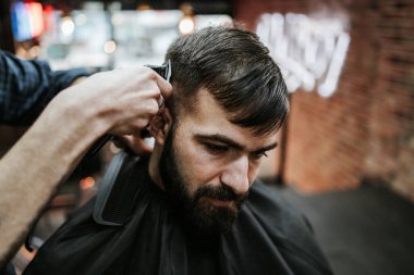 Yakışıklı bir genç adam saç ve sakal stili yaptırıyor ve profesyonel kuaför tarafından giydiriliyor..