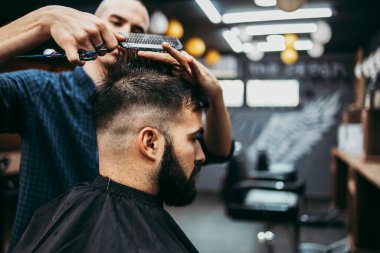 Yakışıklı bir genç adam saç ve sakal stili yaptırıyor ve profesyonel kuaför tarafından giydiriliyor..