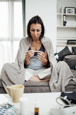 Grip hastası ve yorgun orta yaşlı bir kadın. Evdeki kanepede oturuyor, termometreyi tutuyor ve bej battaniyeye sarılı vücut ısısını ölçüyor..