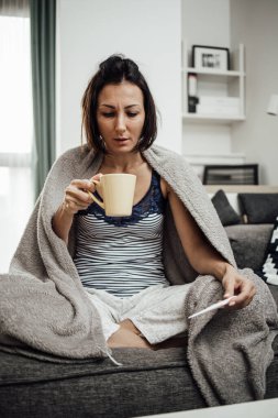 Grip hastası ve yorgun orta yaşlı bir kadın. Evdeki kanepede oturuyor, termometreyi tutuyor ve bej battaniyeye sarılı vücut ısısını ölçüyor..