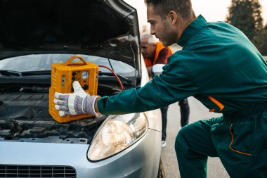 Çekici servisinde çalışan iki yol asistanı atlama marşı ve hava kompresörlü enerji istasyonuyla araba motorunu çalıştırmaya çalışıyor. Yol kenarı yardım kavramı.