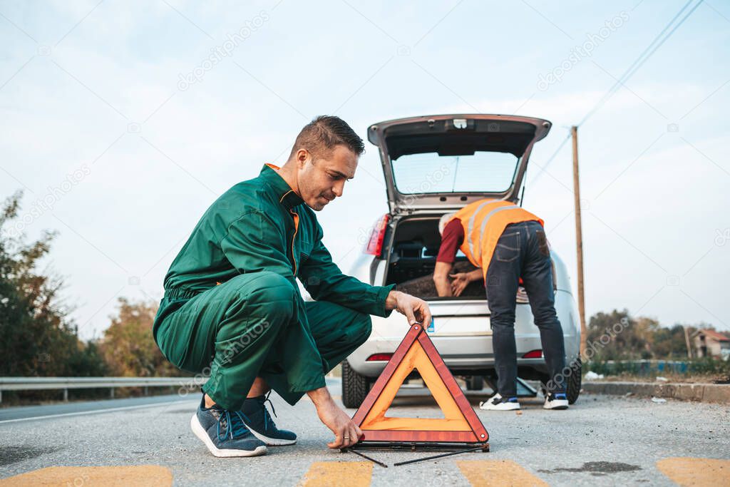 Two road assistant workers in towing service trying to fix car engine. One worker in foreground placing safety triangle on the road while another one doing something in background. Roadside assistance concept.
