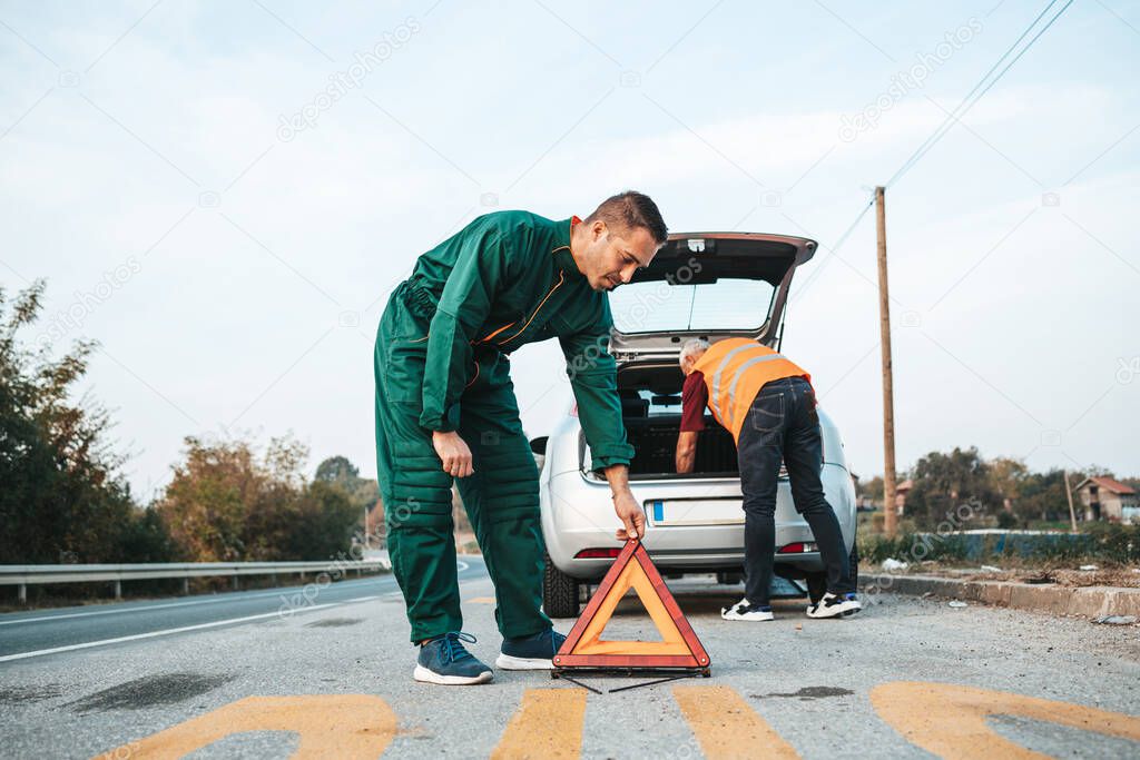 Two road assistant workers in towing service trying to fix car engine. One worker in foreground placing safety triangle on the road while another one doing something in background. Roadside assistance concept.