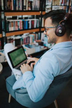 Genç çekici bir adam modern kitapçıda oturmuş, kulaklıkla müzik dinlerken dizüstü bilgisayarını ya da bilgisayarını okuyor ve kullanıyor.