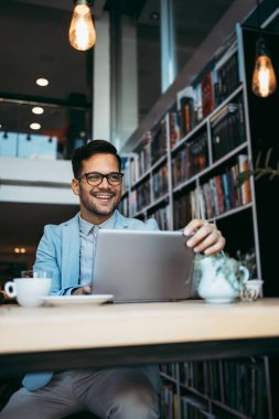 Genç çekici bir adam modern kitapçıda oturuyor, dizüstü bilgisayarını okuyor ve kullanıyor..