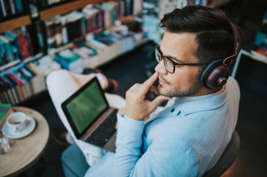 Genç çekici bir adam modern kitapçıda oturmuş, kulaklıkla müzik dinlerken dizüstü bilgisayarını ya da bilgisayarını okuyor ve kullanıyor.
