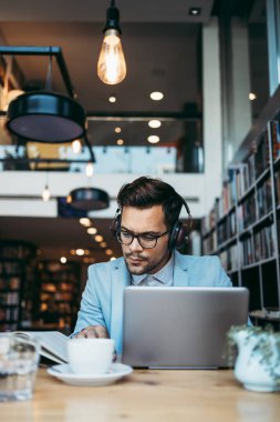 Genç çekici bir adam modern kitapçıda oturmuş, kulaklıkla müzik dinlerken dizüstü bilgisayarını ya da bilgisayarını okuyor ve kullanıyor.