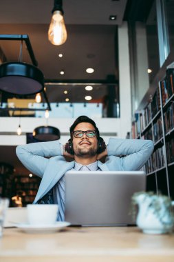 Genç çekici bir adam modern kitapçıda oturmuş, kulaklıkla müzik dinlerken dizüstü bilgisayarını ya da bilgisayarını okuyor ve kullanıyor.