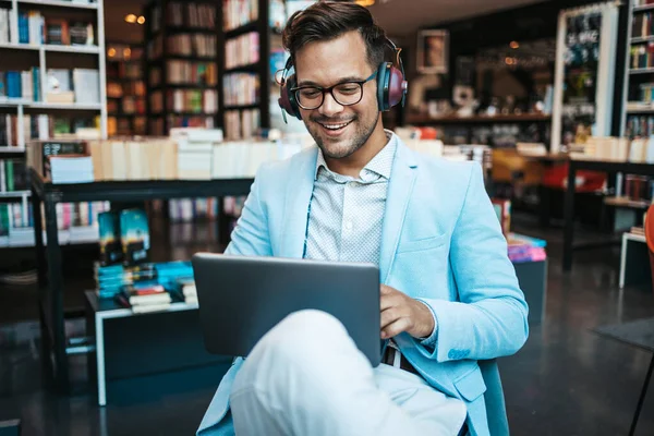Genç çekici bir adam modern kitapçıda oturmuş, kulaklıkla müzik dinlerken dizüstü bilgisayarını ya da bilgisayarını okuyor ve kullanıyor.