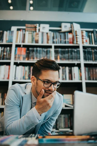 Young nice looking student or writer sitting in college library next to the window and working on his laptop. He is serious and tired. Natural light with strong and deep shadows. Wall with books in the background.