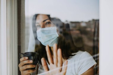 Korkmuş ve endişeli bir kadın dairesinin ya da hastane odasının penceresinden dışarı bakar. Tehlikeli ve bulaşıcı bir virüs korkusuyla yüzüne koruyucu bir maske takıyor. Karantina ve sosyal uzaklık kavramı.