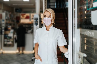 Young and attractive female pharmacist with face protective mask standing at open drugstore doors and looking outside. She is confident and serious. Covid-19 open for business concept. clipart