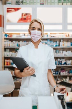 Eczanede çalışan yüzü koruyucu maskeli genç ve çekici bir kadın eczacı. Kendinden emin ve ciddi. Covid-19 iş konseptine açık.